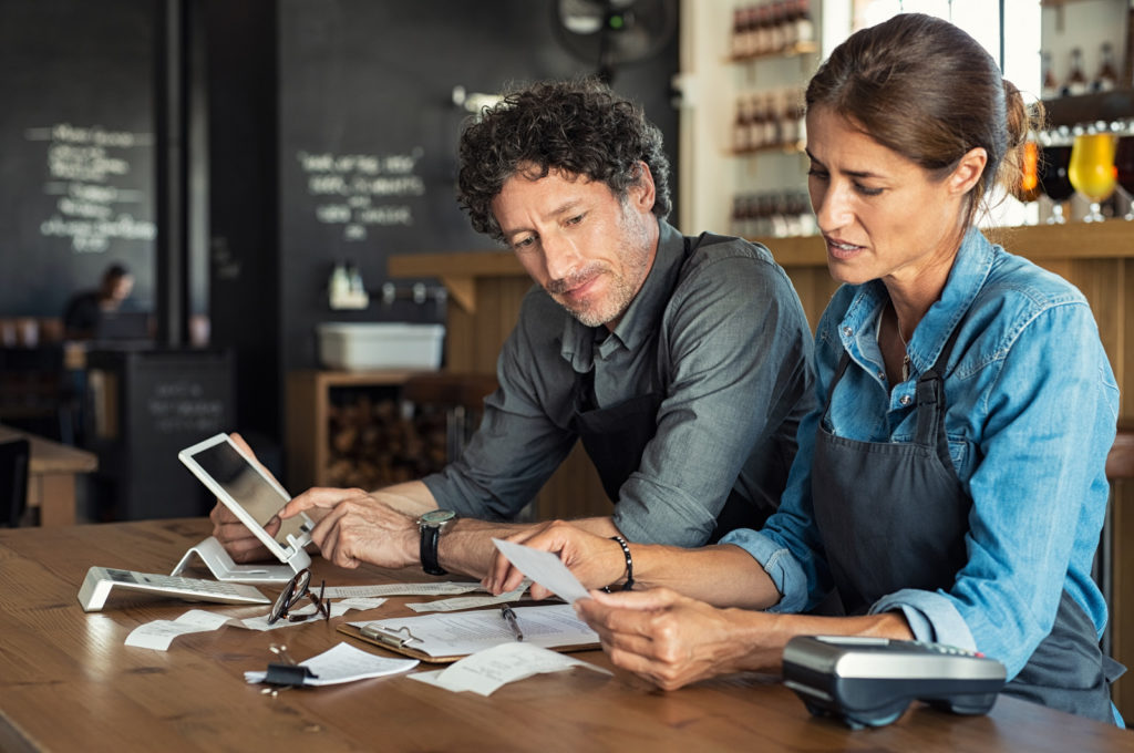 Man and woman sitting in cafeteria discussing finance for the month. Stressed couple looking at bills sitting in restaurant wearing uniform apron. Café staff sitting together looking at expenses and bills.