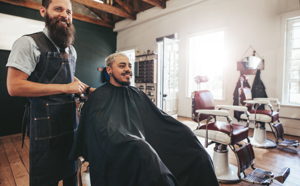 Hairstylist with client sitting at salon and smiling. Hipster man getting haircut at barber shop.