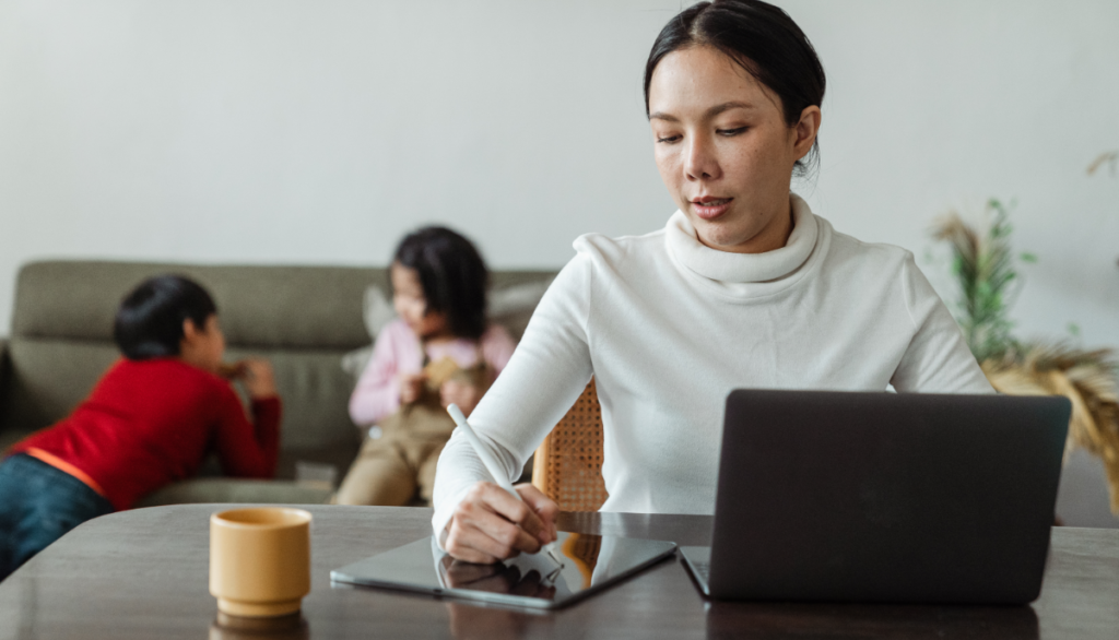 Young Asian mom wearing a white mock neck sweater working on her business from home with her two kids in the background