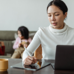 Young Asian mom wearing a white mock neck sweater working on her business from home with her two kids in the background